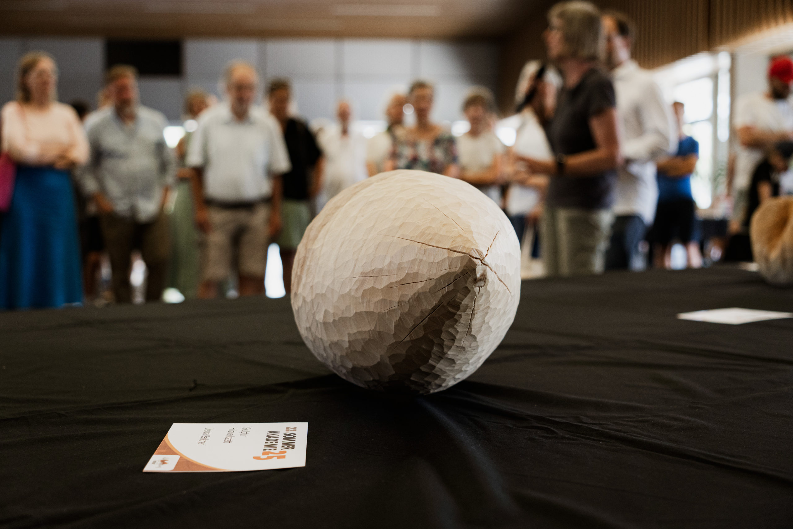 Auf dem Bild ist im Vordergrund eine eiförmige Holzskultptur auf einem Tisch zu sehen.