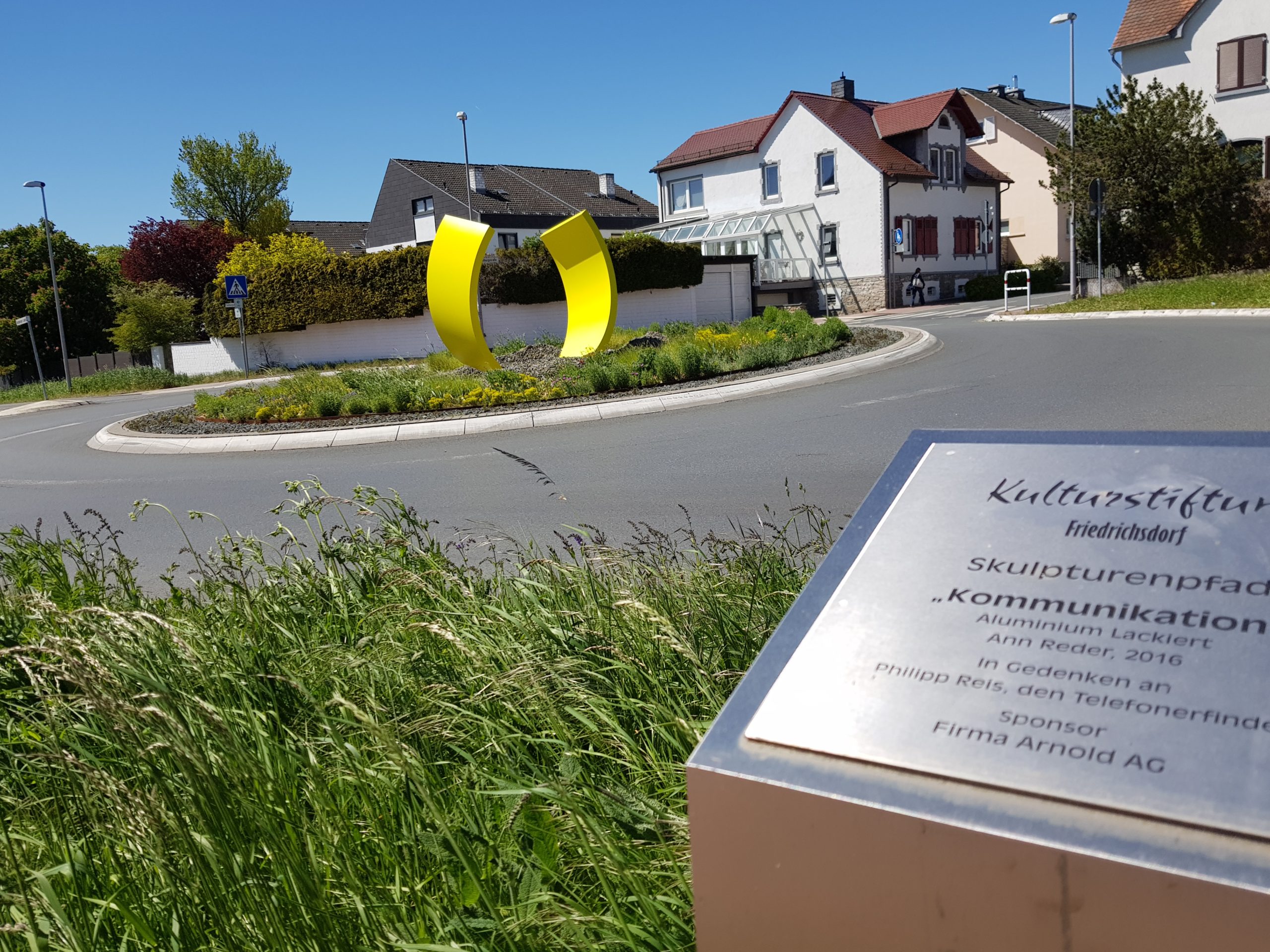 Auf dem Bild ist die gelbe Skulptur Kommunikation auf dem gegrünten Verkehrskreisel zu sehen.