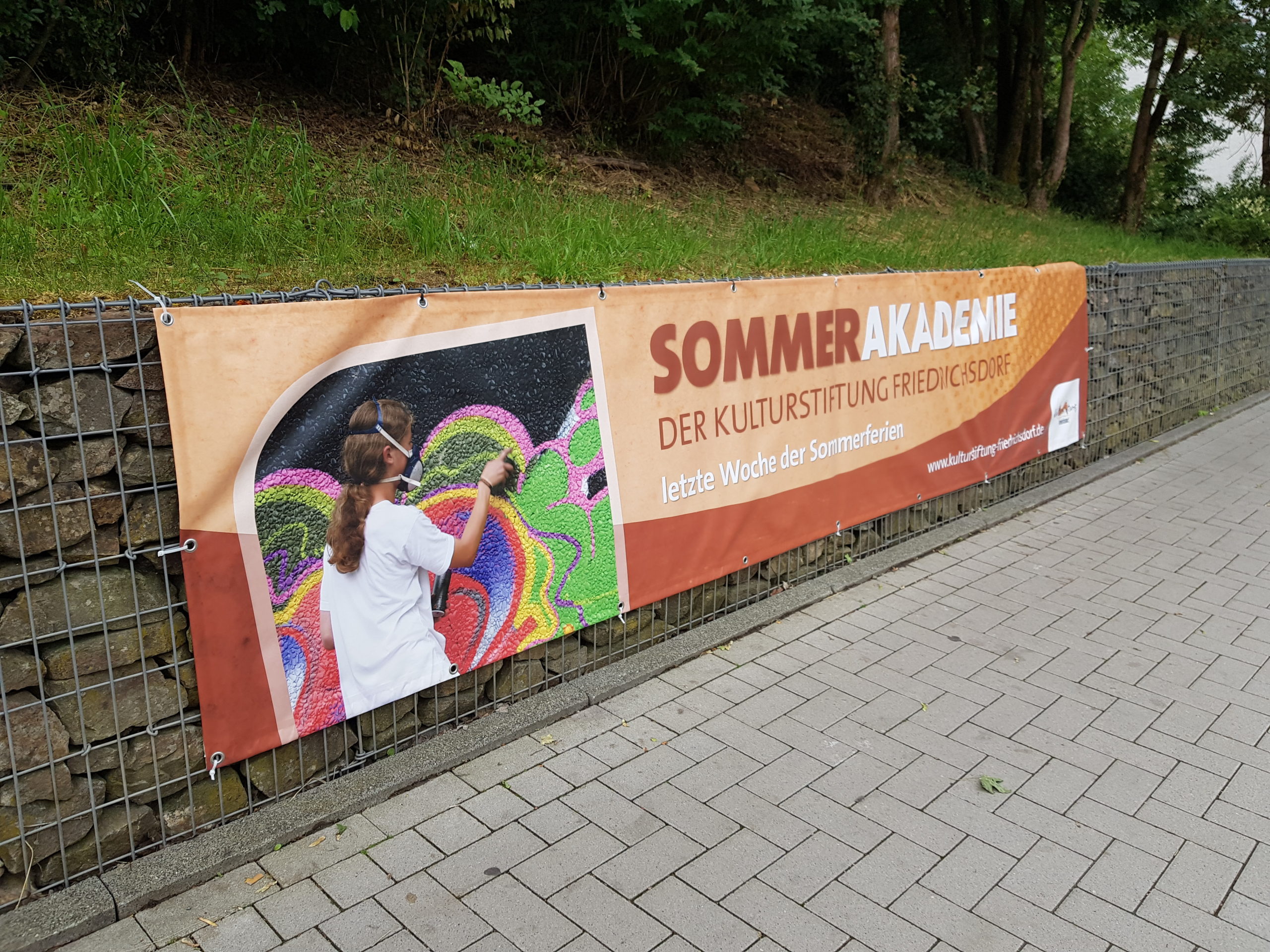 Banner Sommerakademie Auf dem Bild ist das Banner der Sommerakademie an einer Gabionenwand hängend zu sehen.